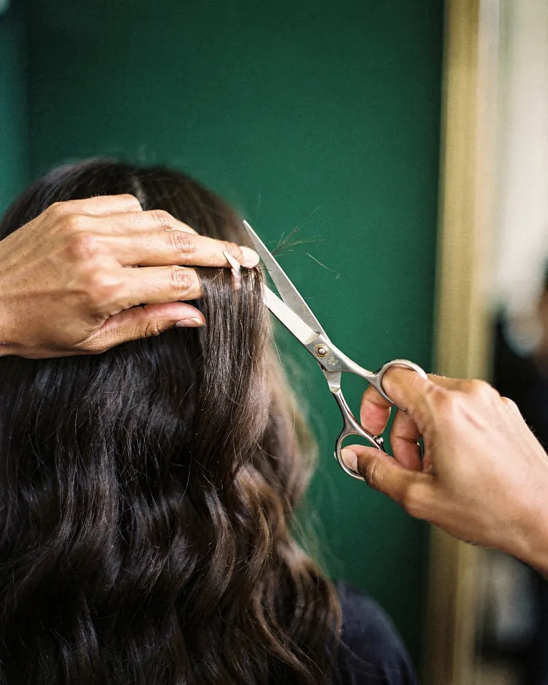 A stylist's hands hold a section of long dark hair and trim the ends with scissors.
